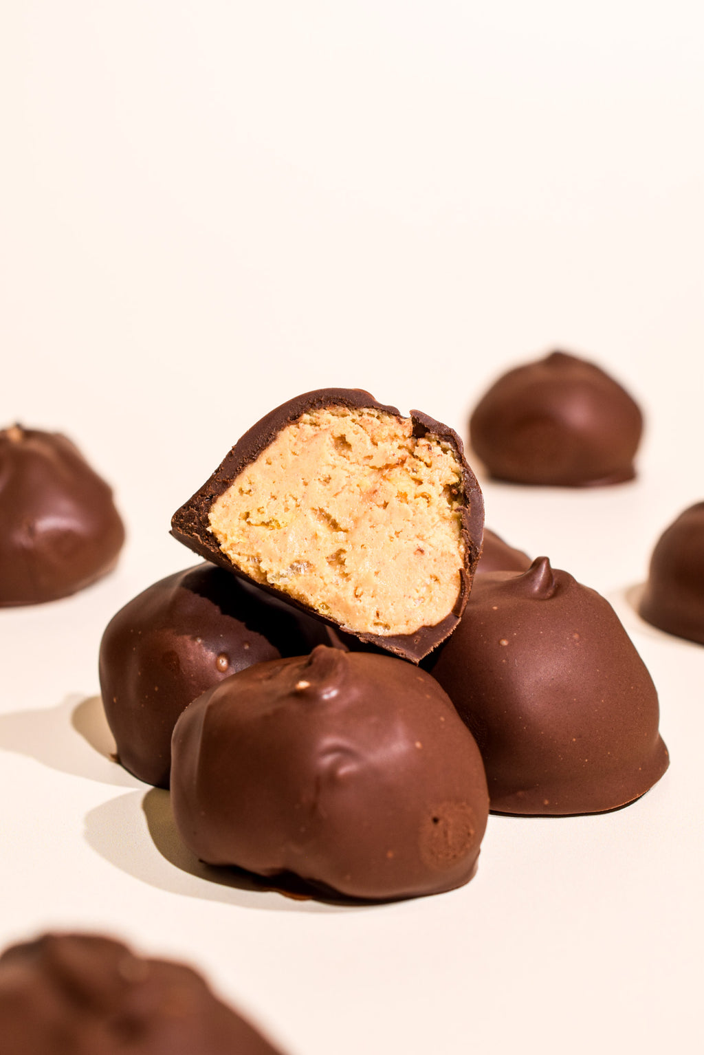 A close-up studio shot of several chocolate-covered candies or chocolates on a white background, with one piece broken open to show a light tan nut or cookie filling.