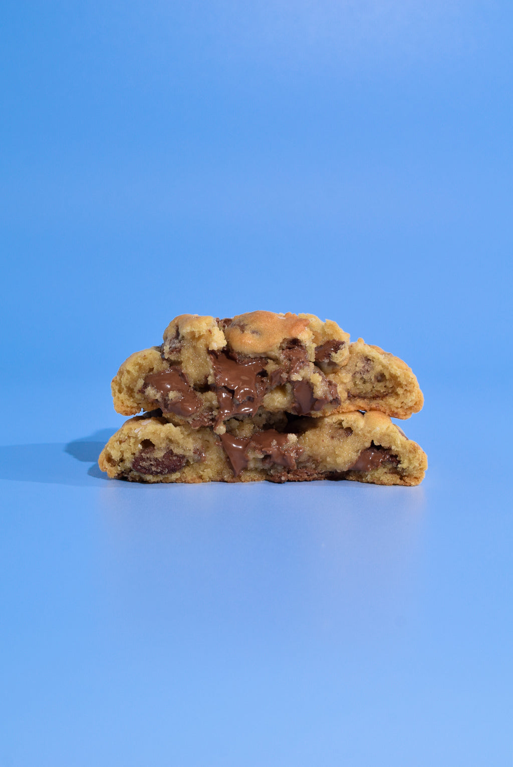 Two halves of a chocolate chip cookie stacked on top of each other, showing melted chocolate chips and cookie dough, set against a solid blue background.