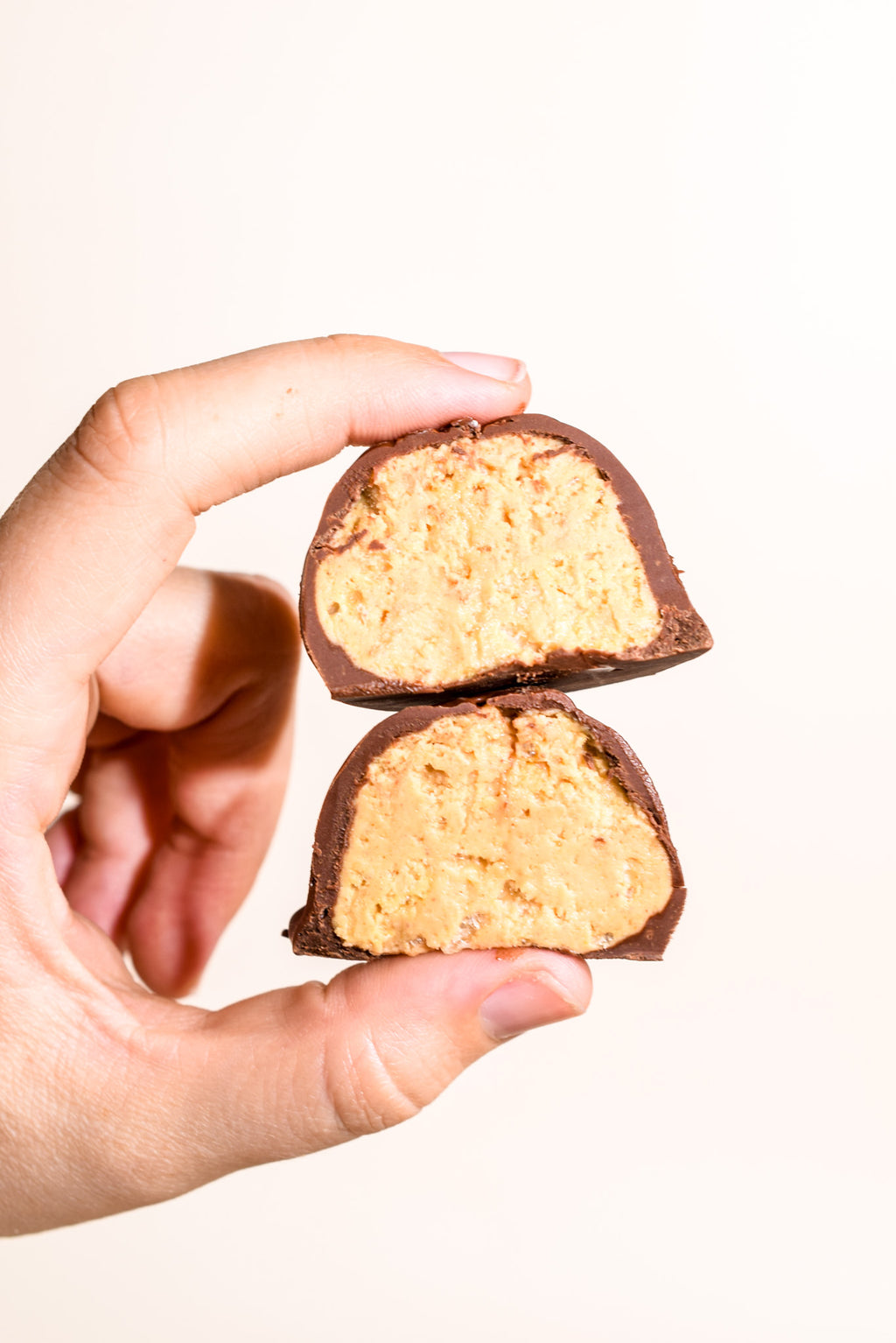 A hand holding two halves of a chocolate-covered peanut butter candy against a plain off-white background.
