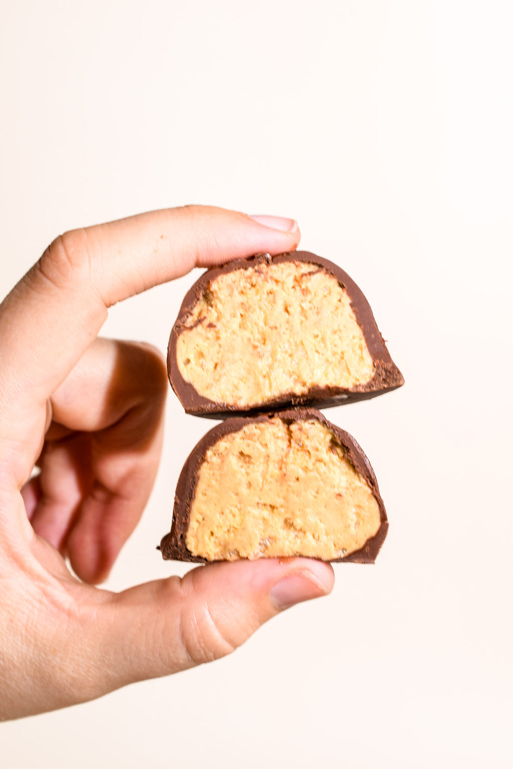A hand holding two halves of a chocolate-covered peanut butter candy against a plain off-white background.