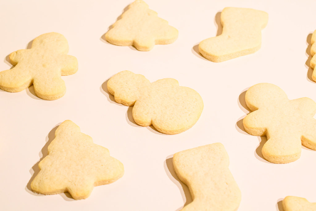 A close-up shot of several pale golden-brown sugar cookies cut into festive shapes, including gingerbread men, Christmas trees, and stockings, arranged on a light cream background.