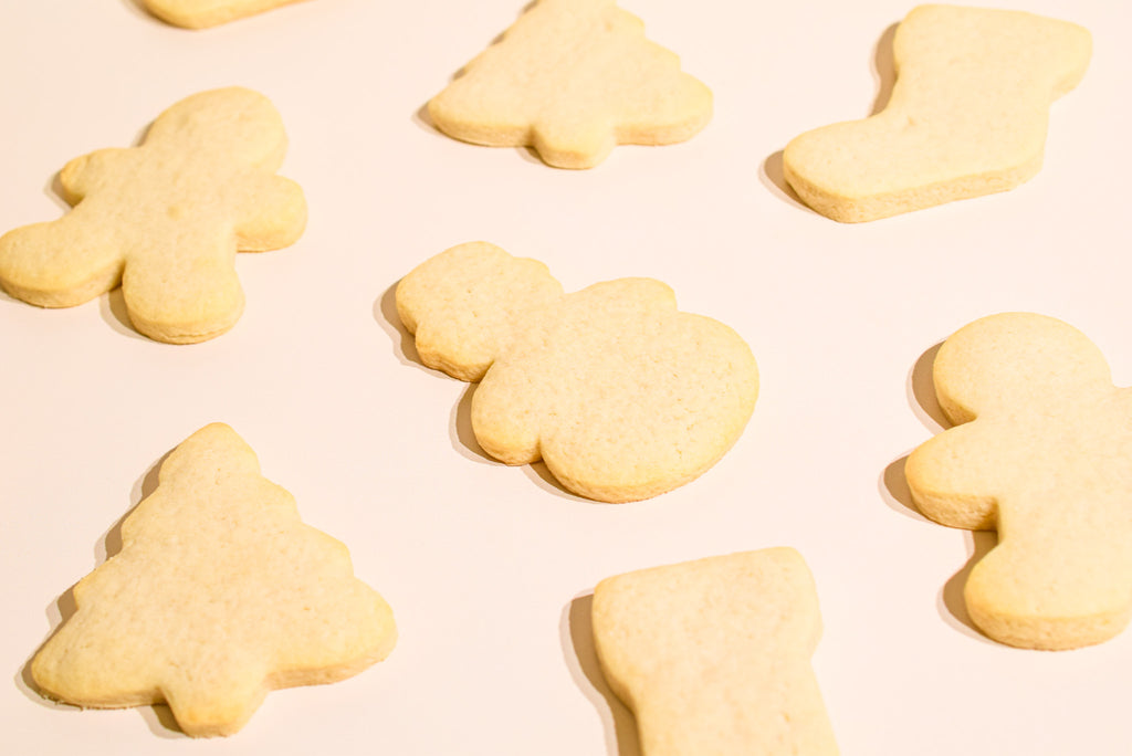 Several pale beige sugar cookies shaped like a Christmas tree, stocking, gingerbread man, and snowman scattered on a plain light background.