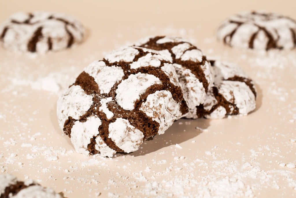 Chocolate crinkle cookies dusted with powdered sugar, arranged on a beige surface with a few more cookies blurred in the background.