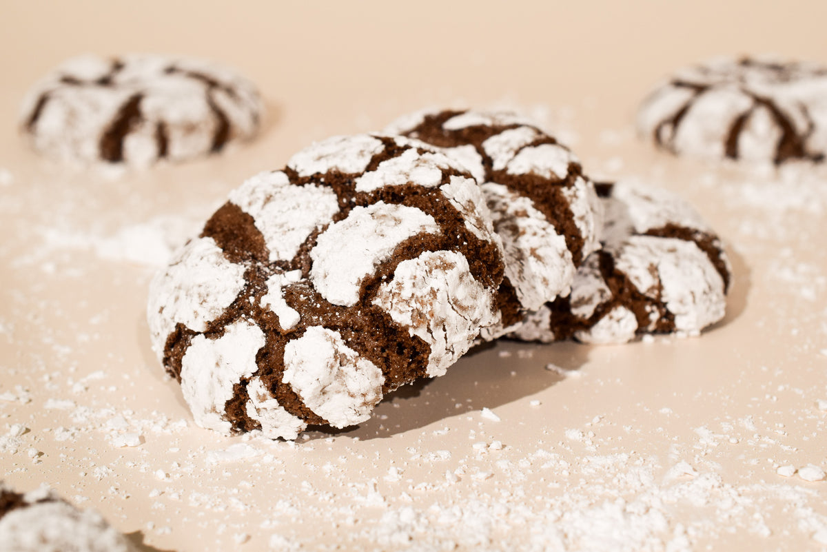 A close-up of a chocolate crinkle cookie dusted with powdered sugar, with two additional cookies blurred in the background on a warm beige surface.