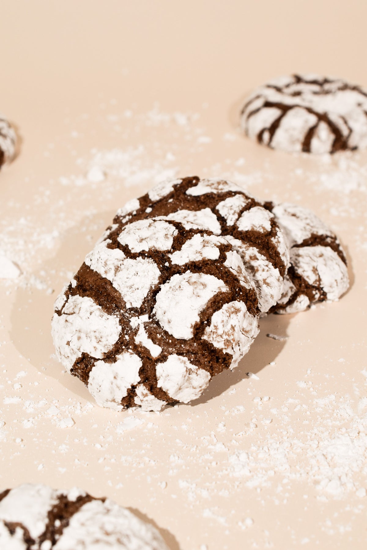 Chocolate crinkle cookies dusted with powdered sugar, arranged on a warm beige surface with more cookies partially visible in the background.