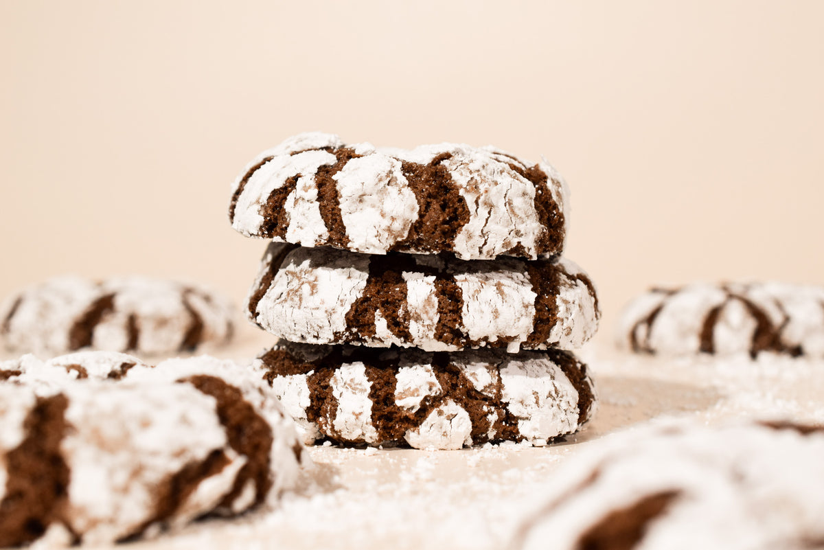 Three stacked chocolate crinkle cookies dusted with powdered sugar, with additional cookies blurred in the foreground and background on a warm beige surface.