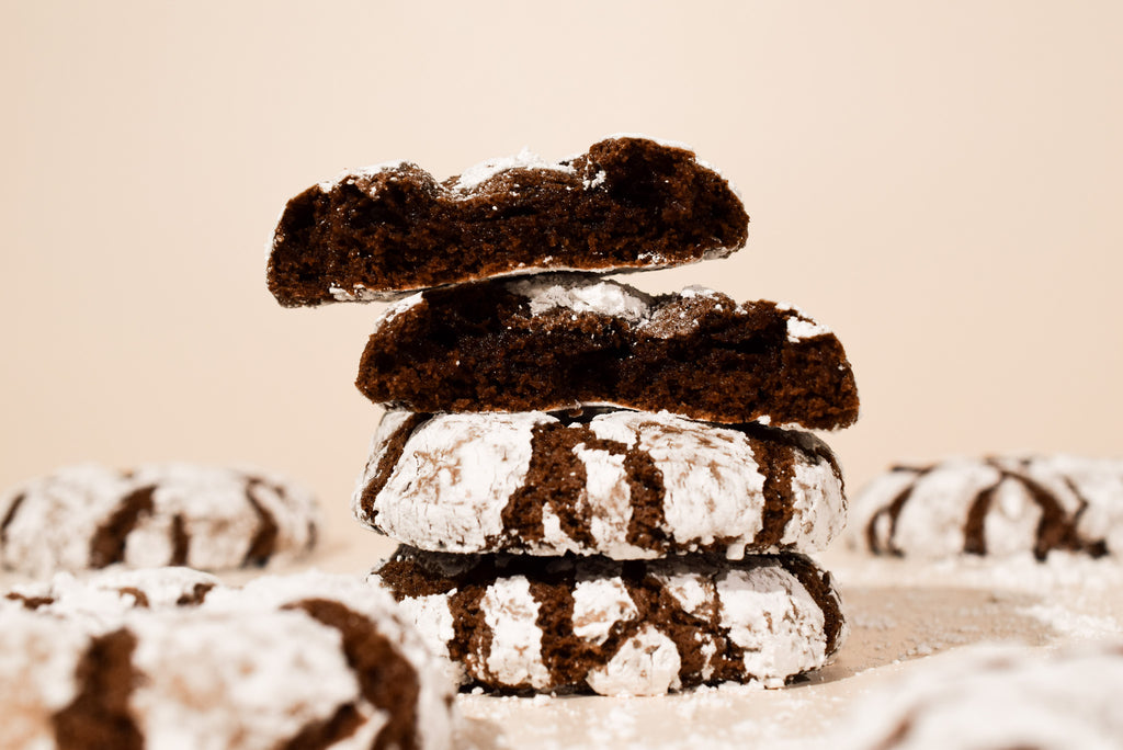 A stack of four chocolate crumb cookies dusted with powdered sugar, photographed on a warm beige background with additional cookies blurred in the foreground and background.