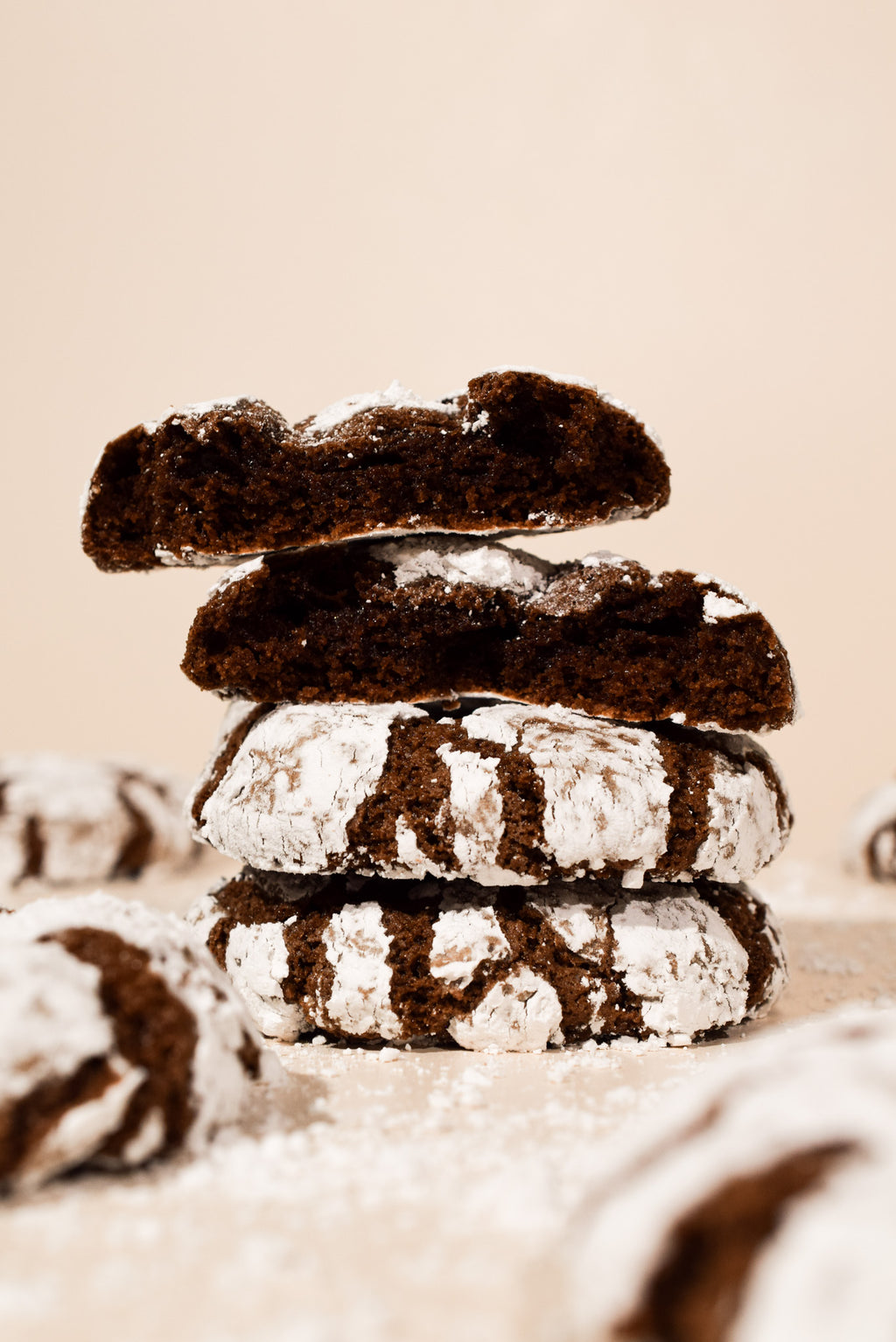 A stack of four chocolate crumb cookies dusted with powdered sugar, photographed on a warm beige background with soft-focus cookies in the foreground and background.