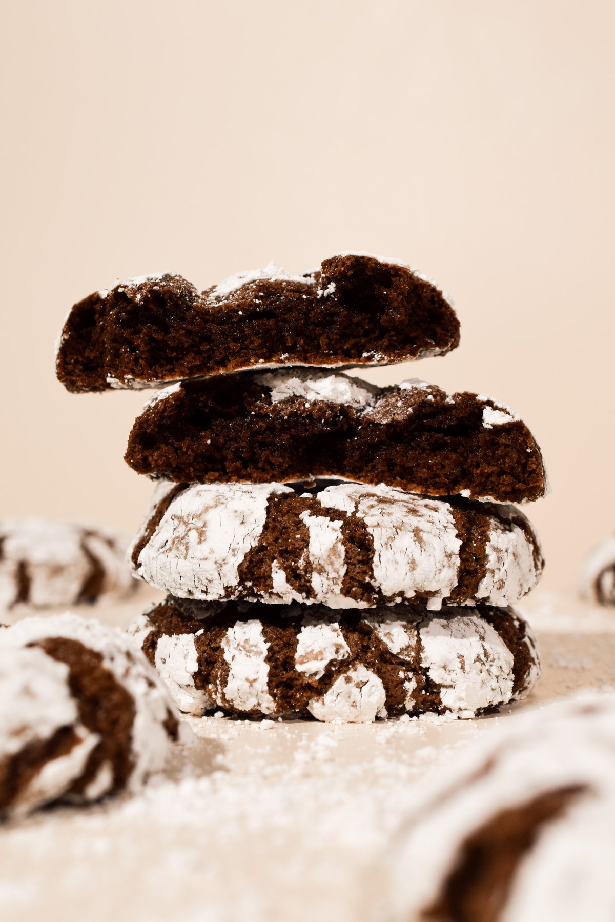 A stack of four chocolate crumb cookies dusted with powdered sugar, photographed on a warm beige background with soft-focus cookies in the foreground and background.