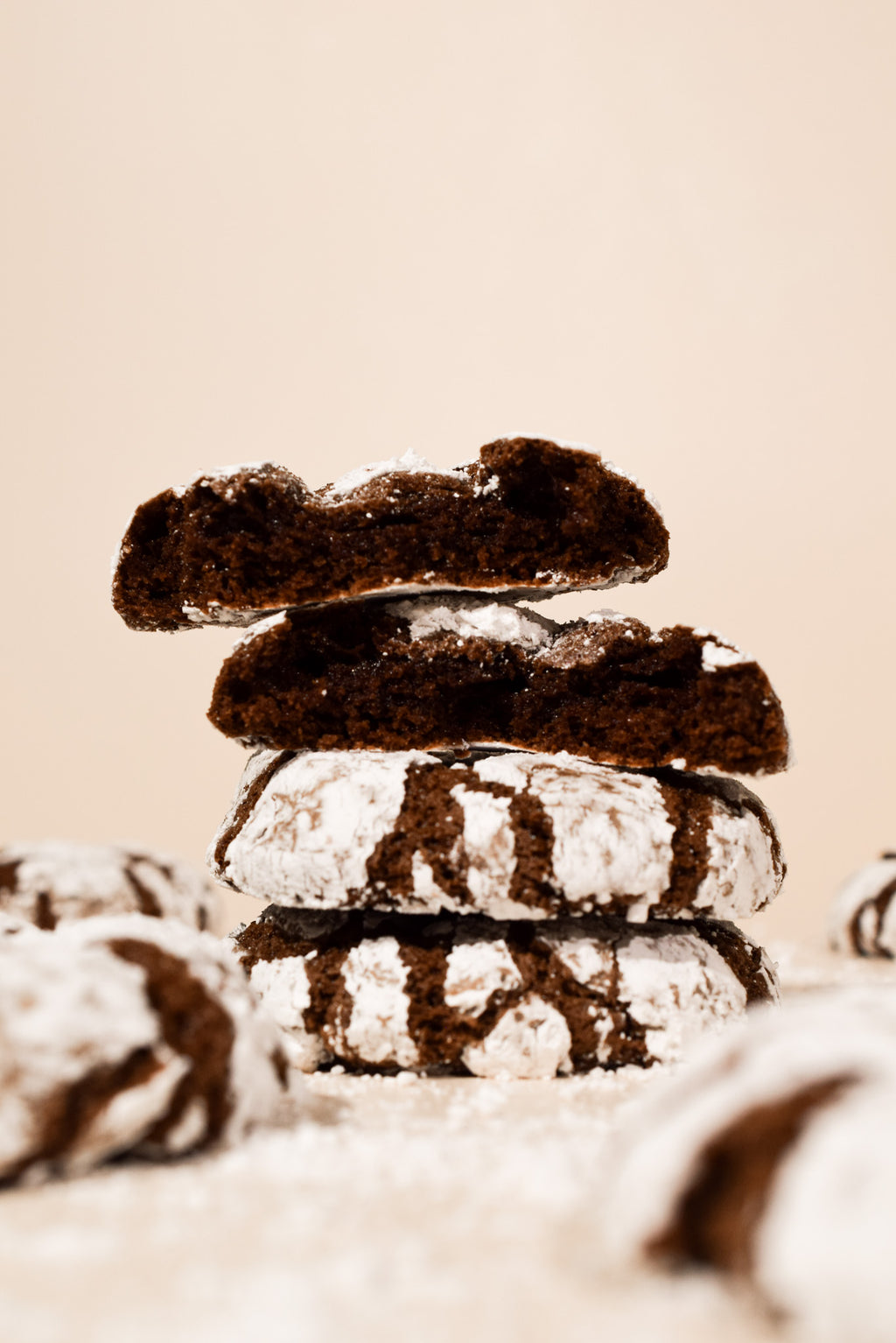 A stack of four chocolate crumb cookies dusted with powdered sugar, photographed on a warm beige background with soft-focus cookies in the foreground.