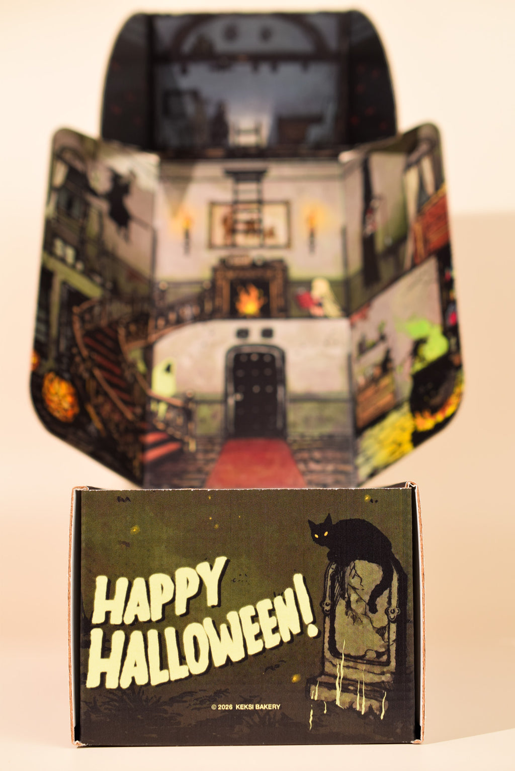 A vintage-style Halloween-themed cardboard display box or packaging piece stands open behind a smaller front panel that reads “Happy Halloween!” with a black cat on a chair illustration. The printed artwork shows a haunted house interior with candles, stairs, and spooky details.