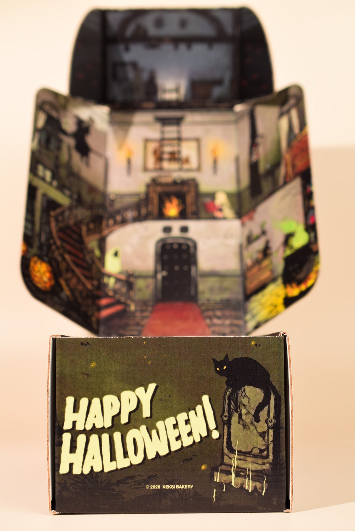 A vintage-style Halloween-themed cardboard display box or packaging piece stands open behind a smaller front panel that reads “Happy Halloween!” with a black cat on a chair illustration. The printed artwork shows a haunted house interior with candles, stairs, and spooky details.