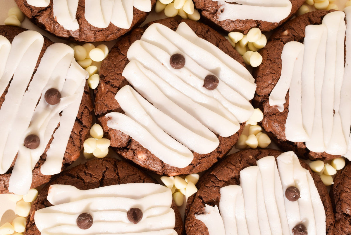 Mummy-themed chocolate chip cookies decorated with white icing, white chocolate chips, and dark chocolate chips, arranged closely together.